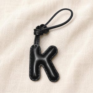 May include: Black leather keychain in the shape of the letter "K". The keychain has a loop for attachment and is stitched around the edges. The background is a light beige fabric.