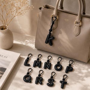May include: A beige leather handbag with gold-coloured hardware is shown with a collection of black letter charms. The letter "K" is attached to the handbag. Additional charms spelling out "LMN OP QRS T" are arranged below.