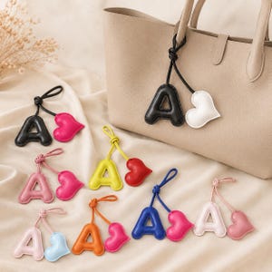 May include: A collection of letter A bag charms, each paired with a small heart charm on a matching cord. The charms come in various colors including black, white, pink, red, yellow, orange, blue, and light pink, attached to a beige tote bag.