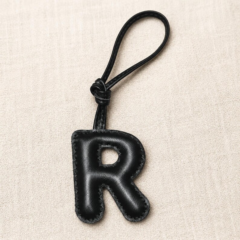 May include: Black leather letter "R" keychain with a loop for hanging. The letter is padded and stitched with a contrasting thread. The keychain is a simple, yet stylish accessory.