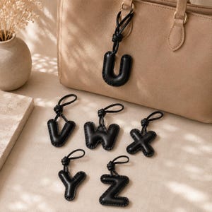 May include: A collection of black leather alphabet letter charms featuring the letters U, V, W, X, Y, and Z. Each letter is padded, stitched along the edges, and attached to a black cord loop for hanging, displayed with a beige handbag.