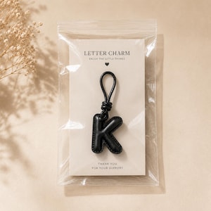 May include: A black leather letter charm in the shape of the letter "K" is displayed in a clear plastic bag. The charm is attached to a black loop. The packaging includes the text "LETTER CHARM" and "ENJOY THE LITTLE THINGS".