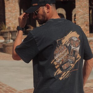 May include: A black short-sleeve t-shirt featuring a graphic print on the back. The design displays two boxing gloves decorated with intricate, swirling patterns in shades of tan, orange, and white, set against a textured tan brushstroke background.