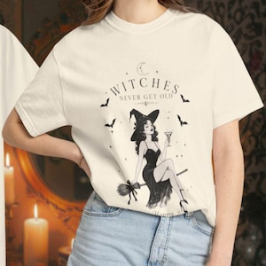 Op de afbeelding: Een ivoorkleurig t-shirt met korte mouwen en een zwarte afbeelding van een heks die op een bezemsteel zit en een martiniglas vasthoudt. De tekst luidt Witches Never Get Old en Happy 30s, omringd door kleine sterren, een maansikkel en vleermuizen.
