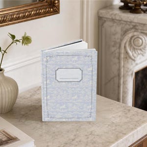 Puede incluir: Un diario de tapa dura en color azul claro con un patrón de toile blanco repetitivo de caballos de carrusel y motivos florales. La portada muestra un marco de etiqueta rectangular central con una línea horizontal para escribir.
