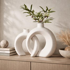May include: Two white ceramic vases with a modern design, one with a circular opening and the other with a curved shape. The larger vase holds a sprig of green leaves with small, dark berries. A decorative home decor item.