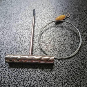 Pode incluir: Uma ferramenta prateada em forma de T com uma pega texturizada e uma ponta preta, juntamente com um laço de arame prateado e uma pequena conta amarela. Os itens estão dispostos sobre uma superfície texturizada cinza escuro.