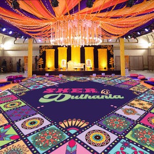 May include: A decorated event space with a stage, seating, and a patterned floor. The ceiling is adorned with orange and purple fabric, lights, and floral garlands. The floor features a large, colorful design with the text "Sher @ Dulhania".