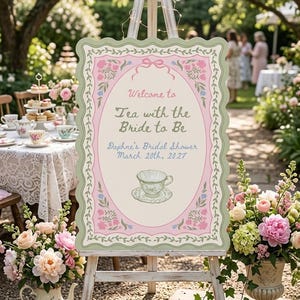 May include: A rectangular sign with a scalloped edge on a wooden easel. The sign features a cream center with pink borders, green floral vines, and a teacup illustration. Text reads: Welcome to Tea with the Bride to Be, Daphne's Bridal Shower, March 20th, 2027.