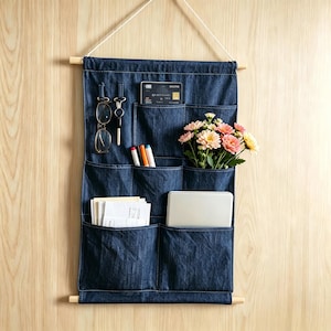 May include: A dark blue denim wall organiser featuring six pockets suspended from a wooden dowel with a white cord. The pockets contain spectacles, a key ring, a black credit card, three markers, a bouquet of pink and yellow flowers, and papers.