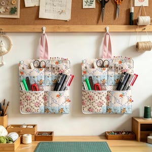 Può includere: Due organizer da parete in tessuto trapuntato identici con un motivo floreale nei toni del rosa, blu e bianco. Ogni organizer presenta diverse tasche riempite con articoli da cucito, tra cui forbici, penne e spilli, e un cinturino rosa.