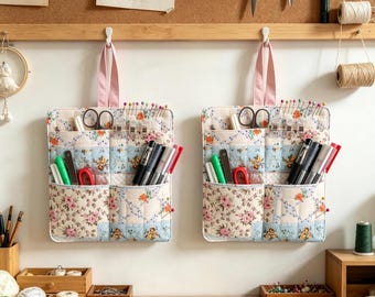May include: Two identical quilted fabric wall organizers featuring a floral pattern in shades of pink, blue, and cream. Each organizer has multiple pockets filled with sewing supplies, including scissors, pens, and pins, and hangs from a pink strap.