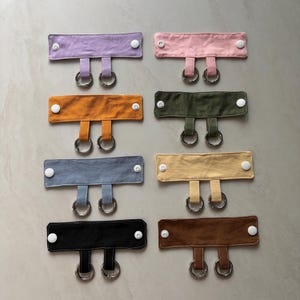 Peut inclure: Huit bandes de tissu rectangulaires disposées en deux colonnes, chacune dotée de deux boutons-pression en plastique blanc aux extrémités et de deux petits anneaux métalliques suspendus au centre par de courtes boucles en tissu, déclinées en lavande, rose, orange, olive, bleu, jaune, noir et marron.