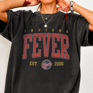 May include: A charcoal grey short-sleeve t-shirt featuring the text INDIANA FEVER in large, distressed red block letters. Below the text, the shirt displays the words EST. 2000 flanking a circular Indiana Fever team logo in red, blue, and gold.