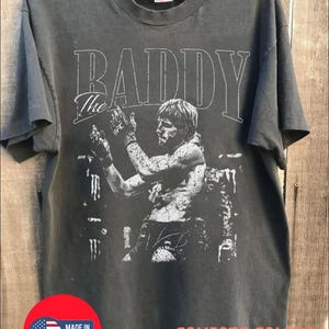 May include: A charcoal gray short-sleeve t-shirt featuring a white graphic print of a fighter in a ring with the text "The BADDY" above. The shirt has a crew neckline and a distressed, vintage-style screen print design on the front chest area.