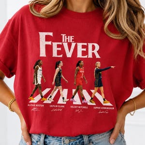 May include: A red short-sleeve t-shirt featuring the text THE FEVER in white distressed lettering. Below the text are four basketball players in uniform with their names Aliyah Boston, Caitlin Clark, Kelsey Mitchell, and Sophie Cunningham printed below.