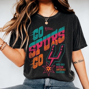 May include: A charcoal gray short-sleeve t-shirt featuring a distressed graphic print that reads GO SPURS GO in teal, pink, and orange. The design includes the NBA Finals logo, the San Antonio Spurs logo, and the text 2025-2026 WESTERN CONFERENCE CHAMPIONS.
