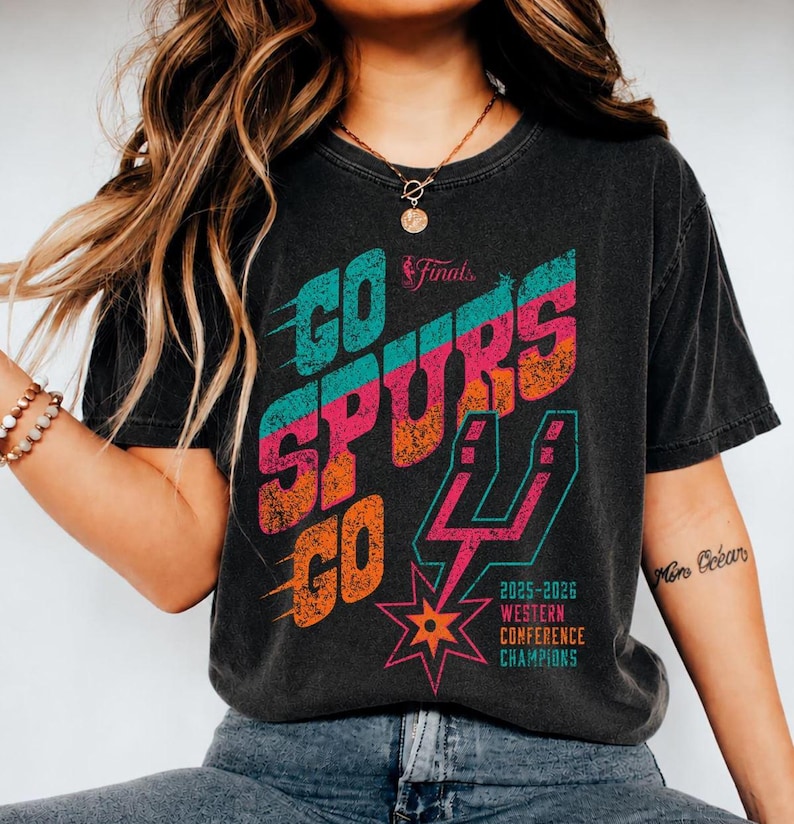May include: A charcoal gray short-sleeve t-shirt featuring a distressed graphic print that reads GO SPURS GO in teal, pink, and orange. The design includes the NBA Finals logo, the San Antonio Spurs logo, and the text 2025-2026 WESTERN CONFERENCE CHAMPIONS.
