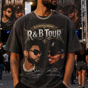 May include: A charcoal gray short-sleeve t-shirt featuring a vintage-style graphic print. The design includes the text "RAYMOND & BROWN R&B TOUR" above portraits of two individuals wearing sunglasses, accompanied by two handwritten-style signatures below.