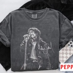 May include: Dark gray t-shirt featuring a black and white image of Michael Jackson performing on stage. The image includes his signature in white. The shirt is folded with a pair of jeans and a phone case with a checkered pattern.