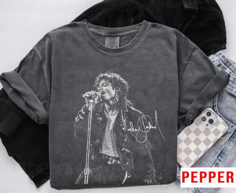 May include: Dark gray t-shirt featuring a black and white image of Michael Jackson performing on stage. The image includes his signature in white. The shirt is folded with a pair of jeans and a phone case with a checkered pattern.