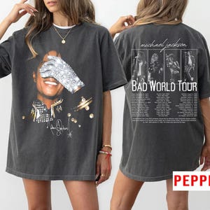 May include: Dark gray t-shirt featuring a graphic of Michael Jackson with a sequined glove covering his eyes. The front of the shirt has his signature and gold accents. The back of the shirt has the text "Michael Jackson Bad World Tour" with tour dates.