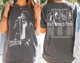 May include: Dark gray t-shirt featuring a black and white image of Michael Jackson on stage, with his signature below. The back of the shirt displays "Bad World Tour" with tour dates and images of Jackson.
