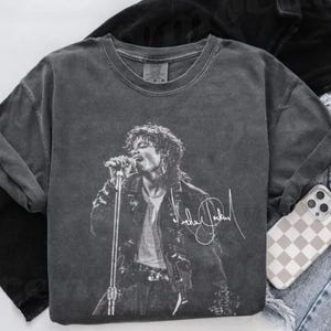 May include: A charcoal gray t-shirt featuring a black and white image of a singer on stage, holding a microphone. The shirt has the singer's signature in white. The shirt is paired with a phone case and denim jeans.