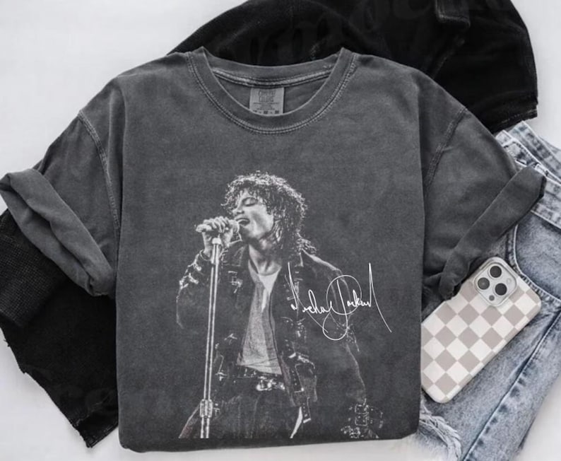 May include: A charcoal gray t-shirt featuring a black and white image of a singer on stage, holding a microphone. The shirt has the singer's signature in white. The shirt is paired with a phone case and denim jeans.