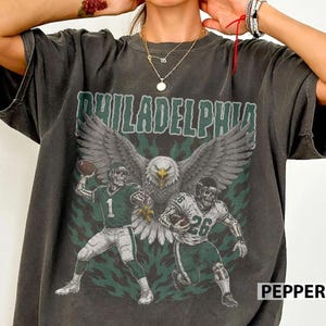 Op de afbeelding: Een antracietgrijs t-shirt met een grafische print van een grote adelaar met gespreide vleugels boven twee skeletachtige footballspelers in shirts met de nummers 1 en 26. Het woord PHILADELPHIA staat in grote groene letters boven de illustratie.