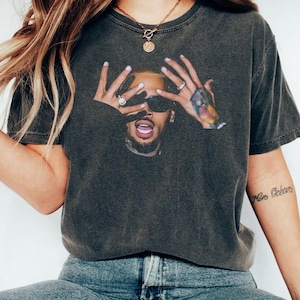 Może przedstawiać: Ciemnoszary t-shirt z krótkim rękawem z nadrukiem graficznym przedstawiającym osobę w ciemnych okularach przeciwsłonecznych, trzymającą dłonie przed twarzą. Osoba ma brodę i otwarte usta.