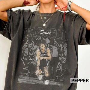 K&ouml;nnte beinhalten: Ein anthrazitfarbenes Kurzarm-T-Shirt mit einem Grafikdruck eines Basketballspielers im Trikot und dem Text VASSELL sowie Devin &uuml;ber dem Bild. Das Design enth&auml;lt einen verblassten Hintergrund mit Zuschauern und eine Signatur links vom Spieler.