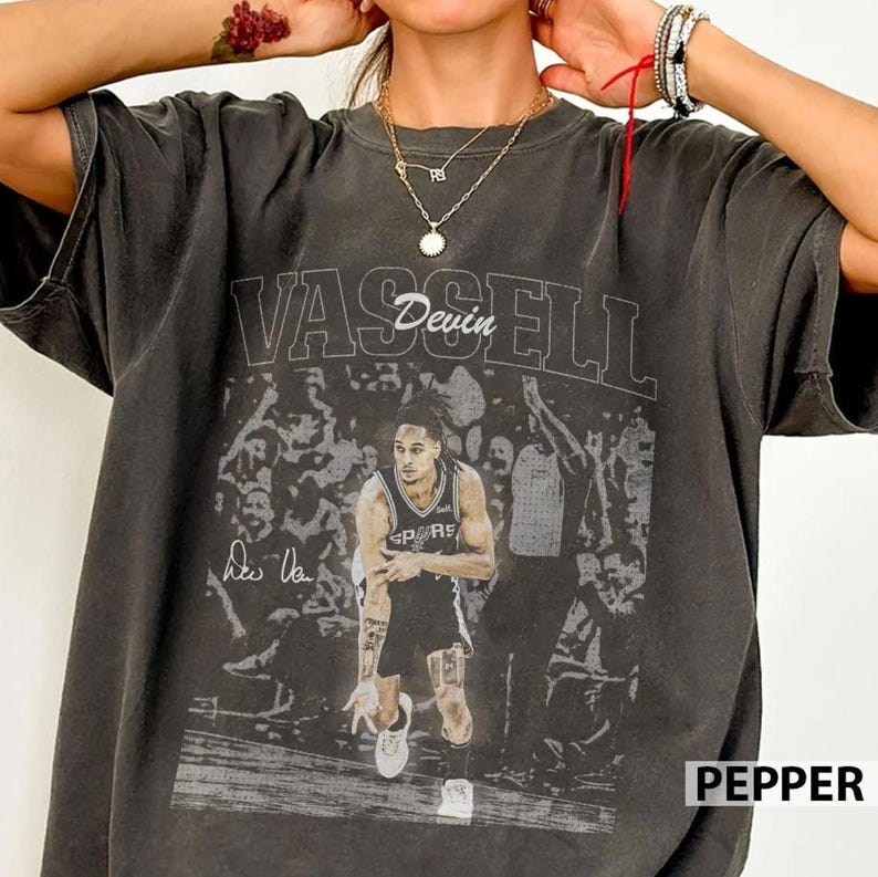 K&ouml;nnte beinhalten: Ein anthrazitfarbenes Kurzarm-T-Shirt mit einem Grafikdruck eines Basketballspielers im Trikot und dem Text VASSELL sowie Devin &uuml;ber dem Bild. Das Design enth&auml;lt einen verblassten Hintergrund mit Zuschauern und eine Signatur links vom Spieler.