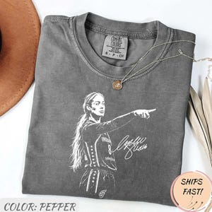 May include: A charcoal gray crew neck t-shirt featuring a white graphic print of a basketball player pointing, with a signature below. The shirt has a Comfort Colors label at the inner neckline and is made of 100% cotton in a size small.