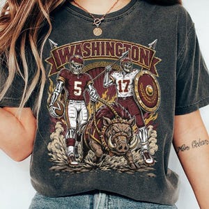 May include: A charcoal grey t-shirt featuring a graphic print with the text WASHINGTON. The design shows two skeleton football players in burgundy and white uniforms, numbered 5 and 17, standing alongside a large, aggressive wild boar.