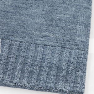 Peut inclure: Un textile en maille bleue présentant une surface texturée avec une bordure côtelée horizontale sur le bas. Le tissu affiche une teinte bleu moyen uniforme avec un aspect doux et légèrement duveteux sur toute la matière tissée.