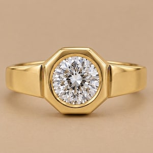 May include: A gold ring featuring a round, brilliant-cut clear gemstone set within an octagonal bezel. The band is smooth and polished, with a wide, solid construction that tapers slightly toward the back, highlighting the central stone.