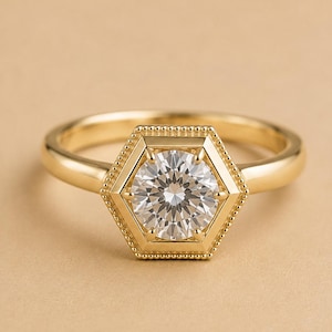 May include: A gold ring featuring a round brilliant-cut clear gemstone set within a hexagonal frame. The hexagonal setting is accented with a delicate beaded texture along its outer edge, and the ring band is smooth, polished, and solid gold.