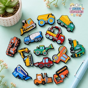 May include: A collection of colorful construction vehicle charms, including dump trucks, excavators, and cement mixers. The charms are in various colors, such as yellow, green, blue, and orange, with black outlines. The charms are scattered on a light blue surface.