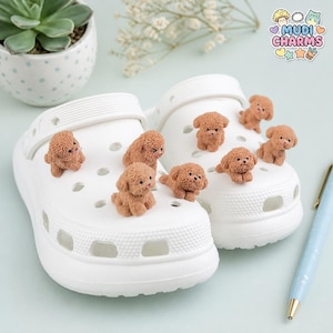 Peut inclure: Une paire de sabots en mousse blanche décorée de six petites breloques texturées en forme de caniche marron. Chaque breloque représente un chien miniature en position assise ou couchée. Les sabots présentent un design perforé.