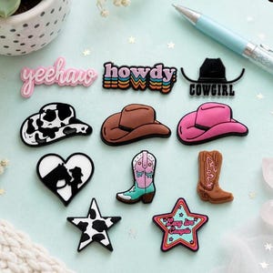 May include: A collection of colorful cowboy-themed charms and accessories. Includes a pink "yeehaw" charm, a "howdy" charm, a black "cowgirl" charm, cowboy hats, boots, a heart with a horse and rider, and a star with "Long live Cowgirls" text.