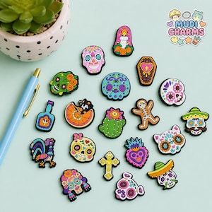 Könnte beinhalten: Eine Sammlung farbenfroher, dekorativer Charms mit Tag der Toten-Themen. Die Charms umfassen Zuckerschädel, einen Sarg, einen Kaktus, einen Lebkuchenmann und einen Hahn. Die Charms befinden sich auf einer hellblauen Oberfläche.