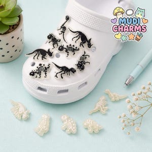 May include: A white foam clog shoe decorated with multiple black dinosaur skeleton charms. Additional loose, off-white dinosaur skeleton charms are arranged nearby. A logo in the corner displays the text MUDI CHARMS with icons.
