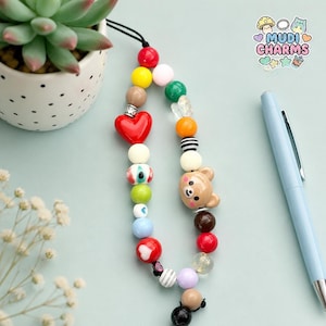 Puede incluir: Un colgante para teléfono con una variedad de cuentas coloridas, incluyendo un corazón rojo, una cara de oso marrón y una cuenta con un diseño de ojo. El colgante está ensartado en un cordón negro e incluye varias cuentas redondas amarillas, verdes, azules y blancas.