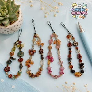 Puede incluir: Cuatro colgantes de teléfono con cuentas con varios diseños, incluyendo mariposas y flores, en tonos naranja, rosa y marrón. Cada colgante tiene un lazo negro y detalles dorados. Los colgantes se muestran sobre una superficie azul claro.