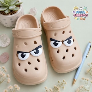 May include: A pair of beige slip-on clogs featuring perforated holes and decorative white and black angry eye charms. The clogs have a heel strap and a smooth finish. A logo in the corner reads MUDI CHARMS with icons of a mushroom, beads, and a drink.