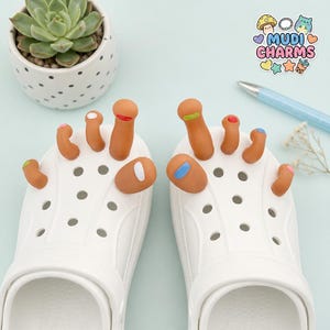 May include: White clogs adorned with brown toe charms, each with a different colored nail polish. A succulent plant in a polka-dotted pot and a pen are in the background. The image also includes a sticker with the text "Mudi Charms".