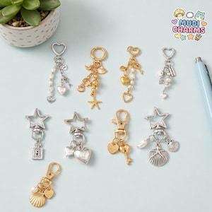 May include: A collection of keychains with various charms, including hearts, stars, seashells, and pearls. The keychains are in gold and silver tones, with lobster claw clasps and heart-shaped key rings. The image includes the text "Mudi Charms".