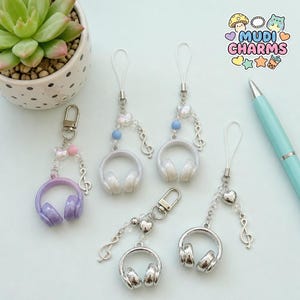 Puede incluir: Cinco colgantes de auriculares con herrajes plateados. Un colgante morado, dos blancos y dos plateados. Cada uno tiene una nota musical y detalles de corazón o cuentas. Los colgantes están unidos a un lazo blanco. El logo "Mudi Charms" es visible.