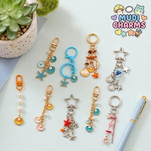 May include: A selection of keyrings with seaside-themed charms, such as seashells, starfish, and pearls. The keyrings have gold, silver, and blue details, with some featuring star-shaped or circular designs. The image also includes the text "Mudi Charms".